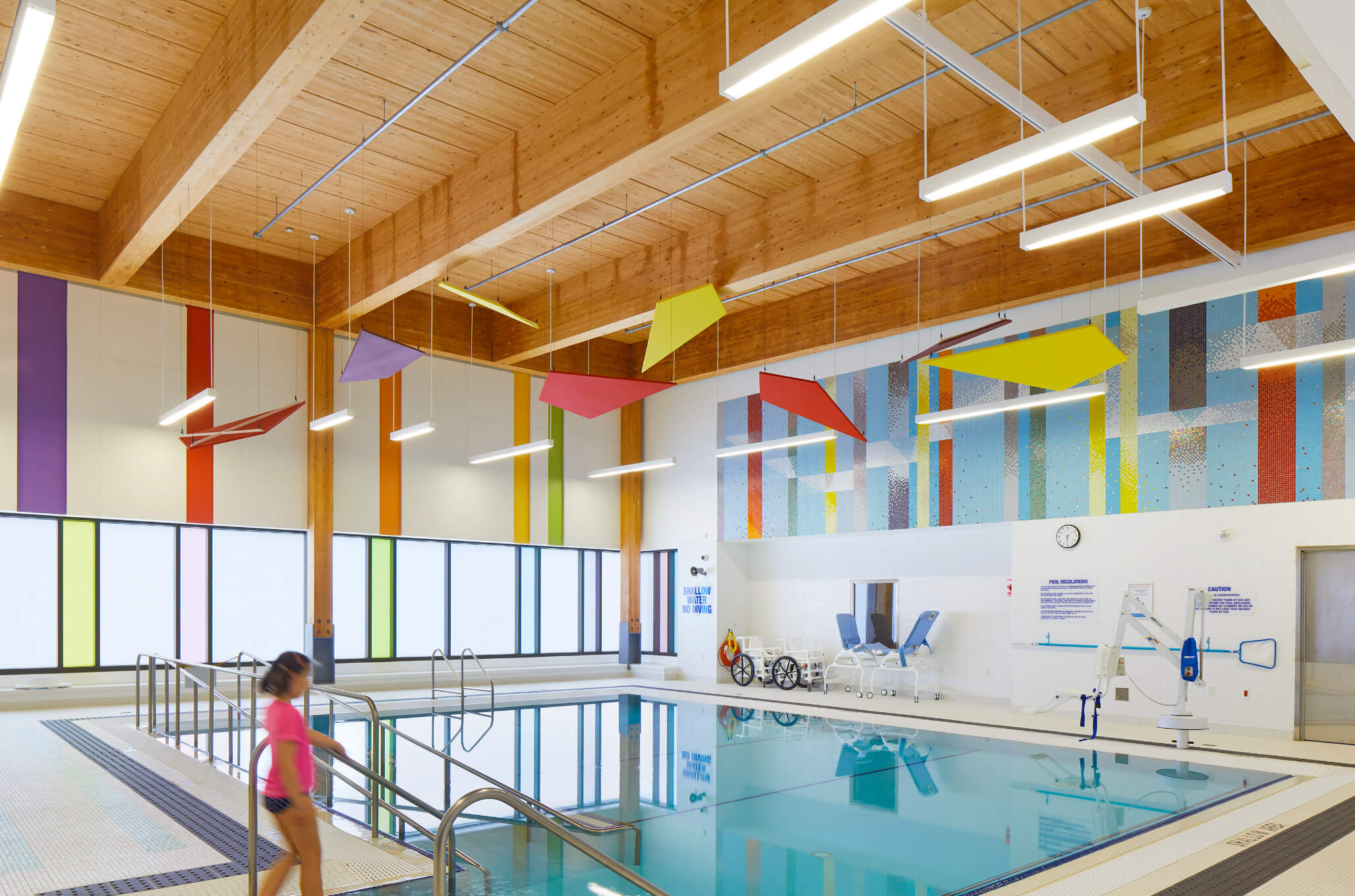Photo of the pool at ErinoakKids Brampton. There is a swimmer in a pink swim shirt about to walk down the stairs and into the pool. On the far side of the pool, along the wall, are accessibility equipment such as water wheelchairs.